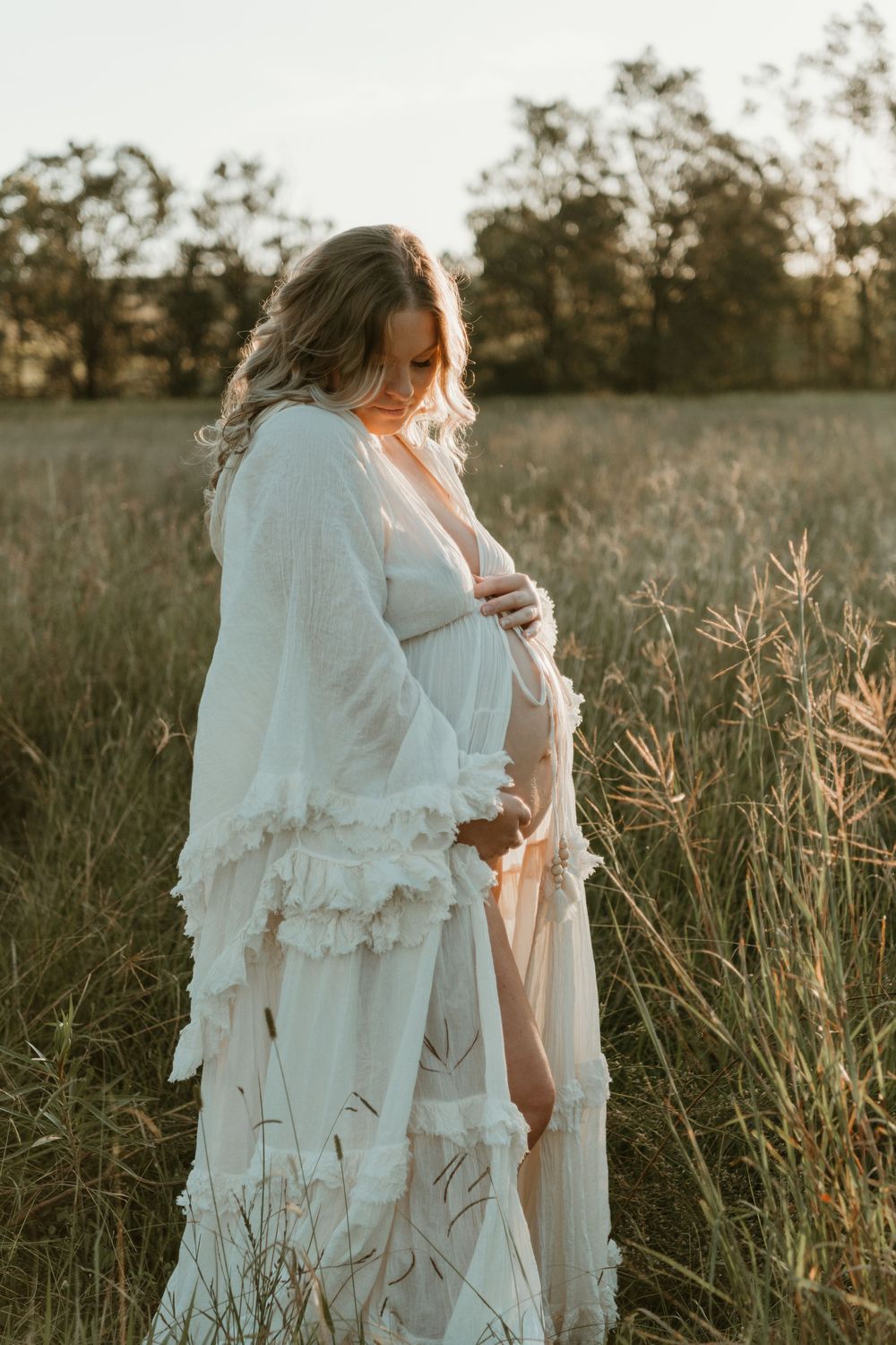 Pregnant woman in white lace robe cradling her baby bump in a golden field at sunset.