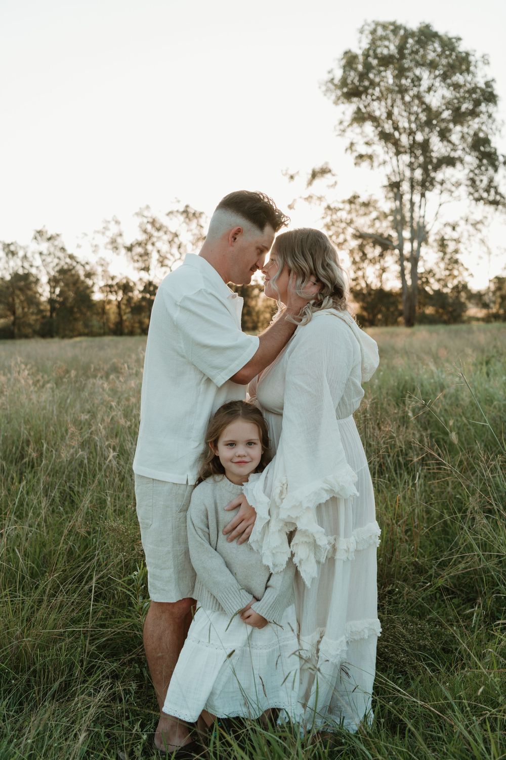 Family of three in white outfits standing in a golden field at sunset, parents touching foreheads with daughter between them.