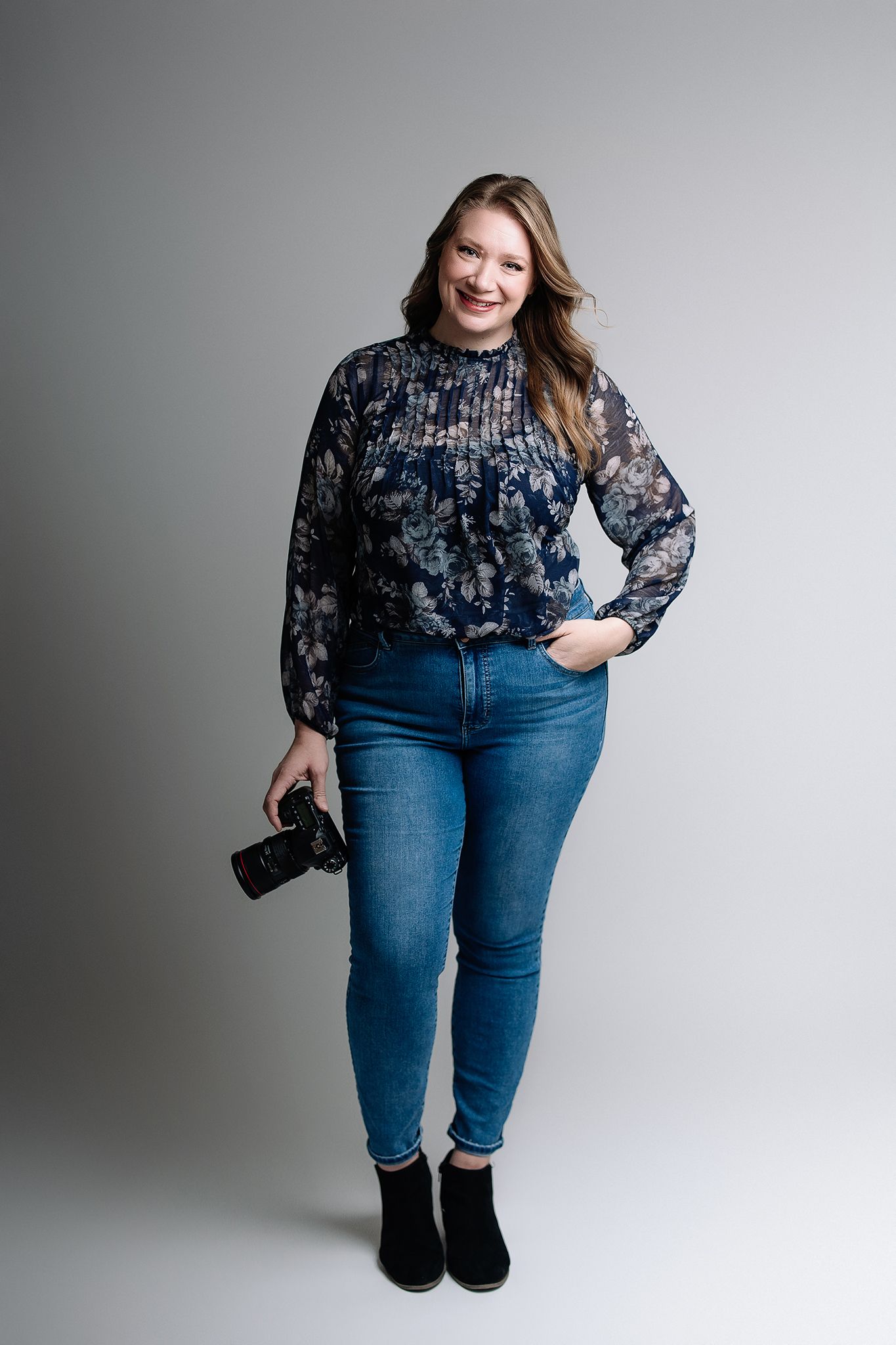 Studio portrait of professional newborn photographer Sarah Savage on white backdrop standing and holding camera.