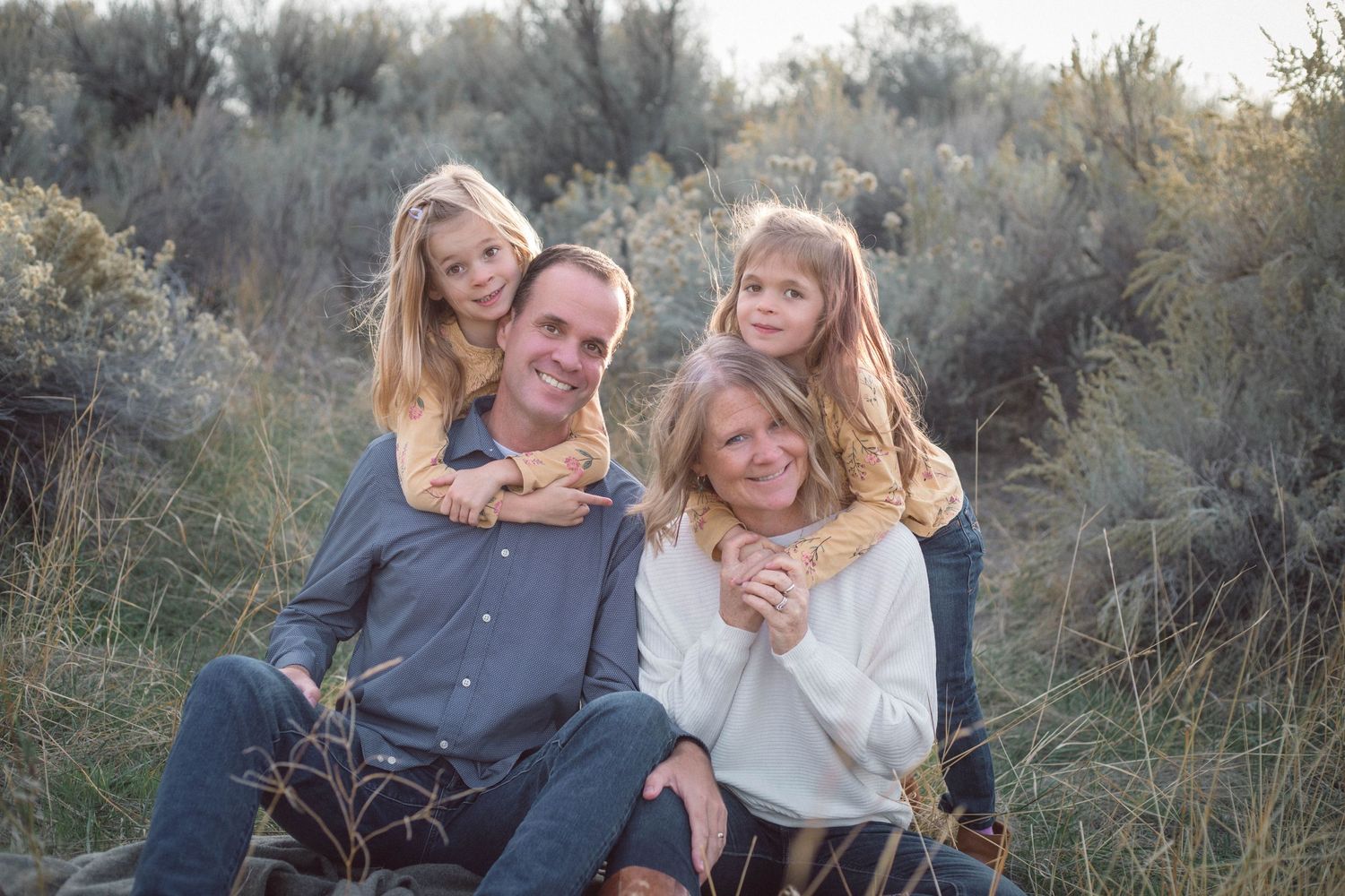 A happy family of four poses together in a natural outdoor setting with sage brush.
