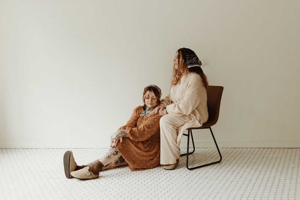 a mother a daughter duo, the daughter leaning on the front of the mothers legs while mother sits in a chair and looks out the window