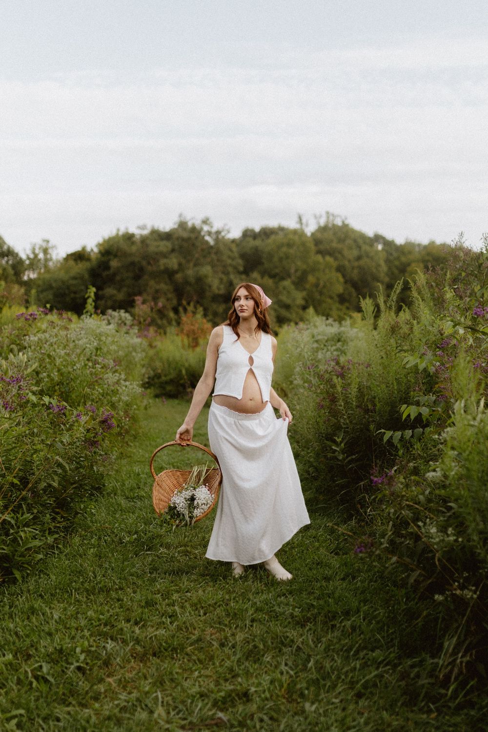 a pregnant woman standing with ankles crossed in a summer field holding a basket of wildflowers