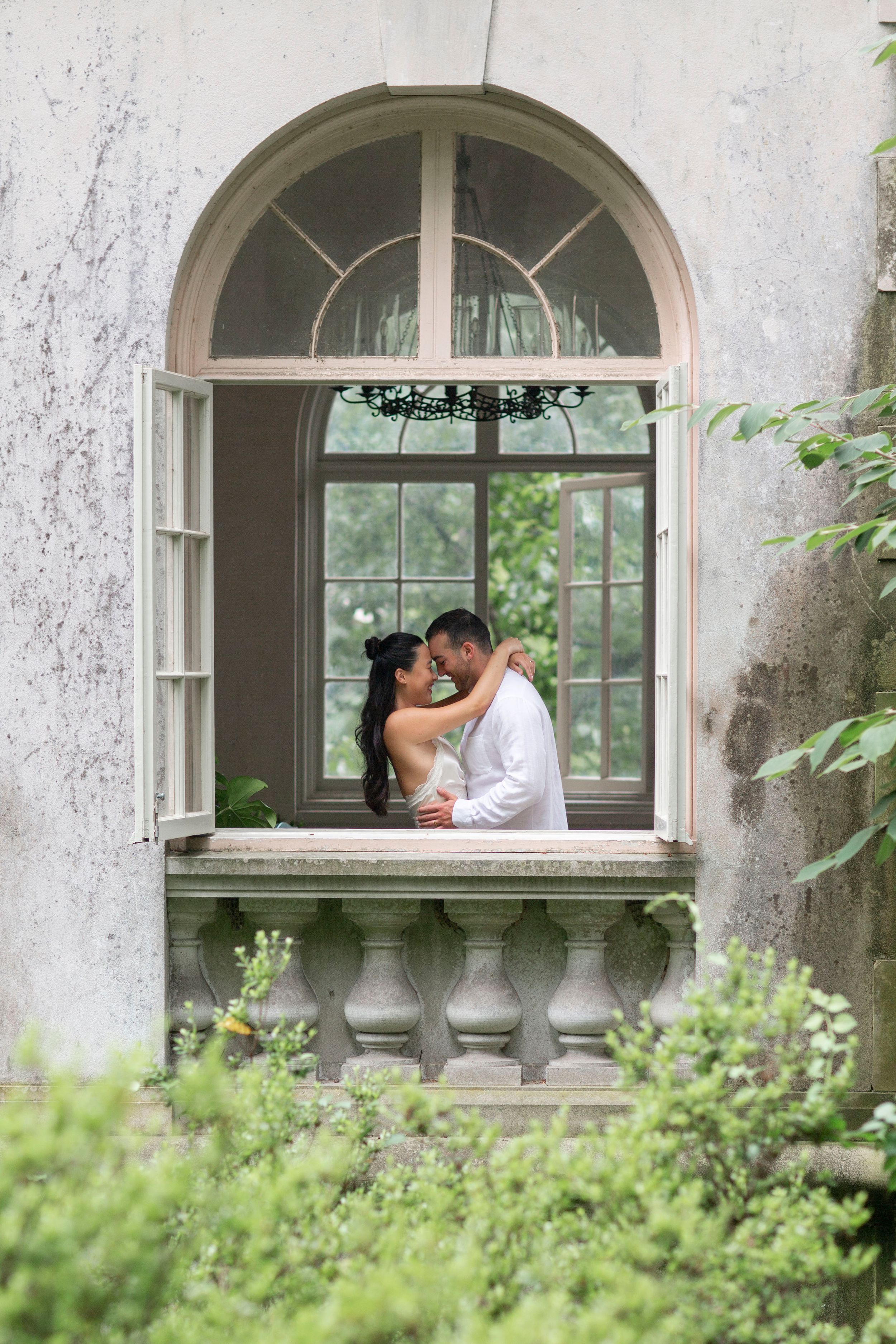 Bride and groom embrace romantically in an arched window of a vintage stone building.