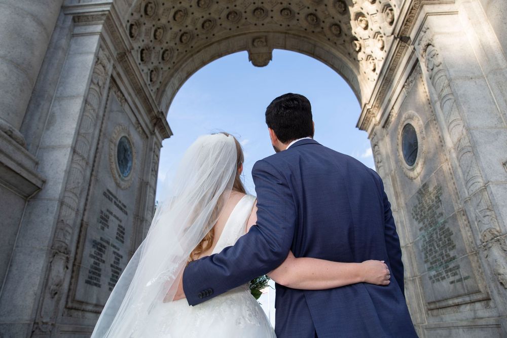 Bride and groom viewed from behind embracing under a grand ornate stone arch.