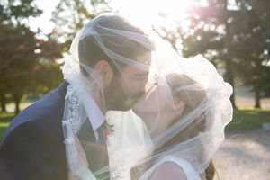 Bride and groom sharing a romantic kiss under a flowing white veil with soft golden sunlight in the background.