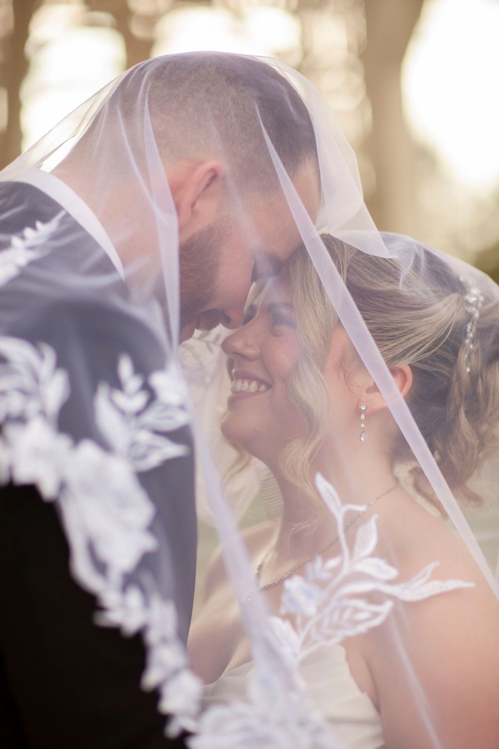Bride and groom sharing an intimate moment under a lace wedding veil in golden sunlight.