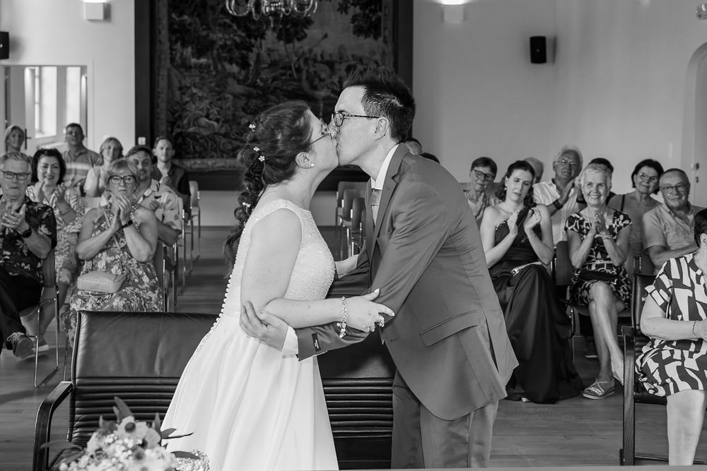 Black and white photo of a couple sharing their first kiss at a wedding ceremony while guests applaud in the background.