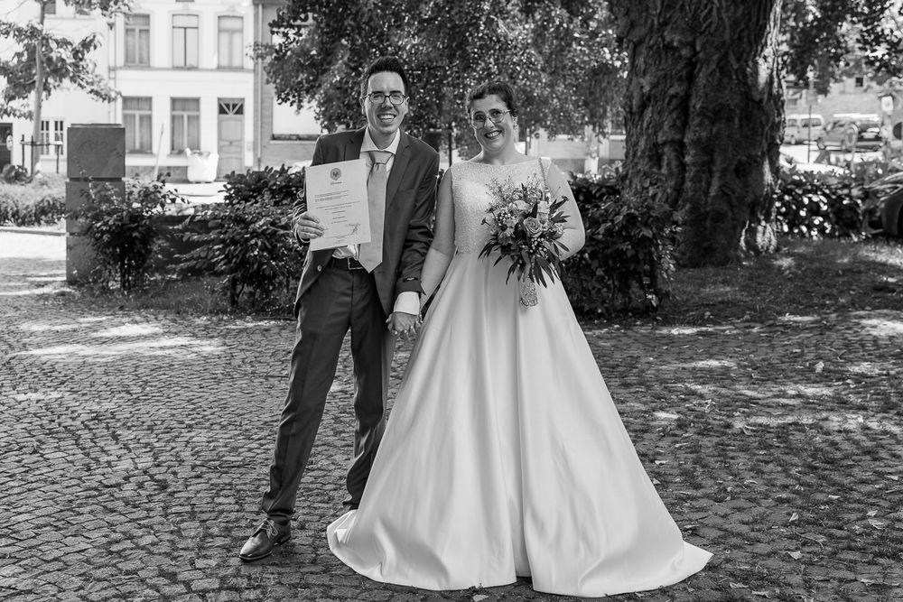 Black and white photo of a smiling bride and groom posing outdoors on their wedding day near a large tree.
