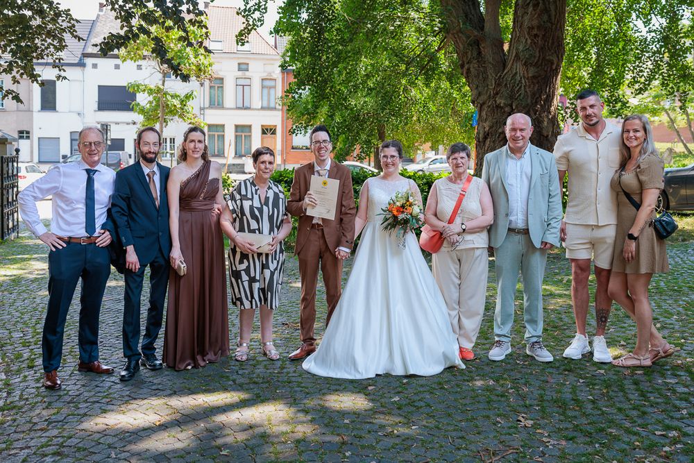 Wedding party posing outdoors near a large tree, bride in white gown holding colorful bouquet with guests in formal attire.