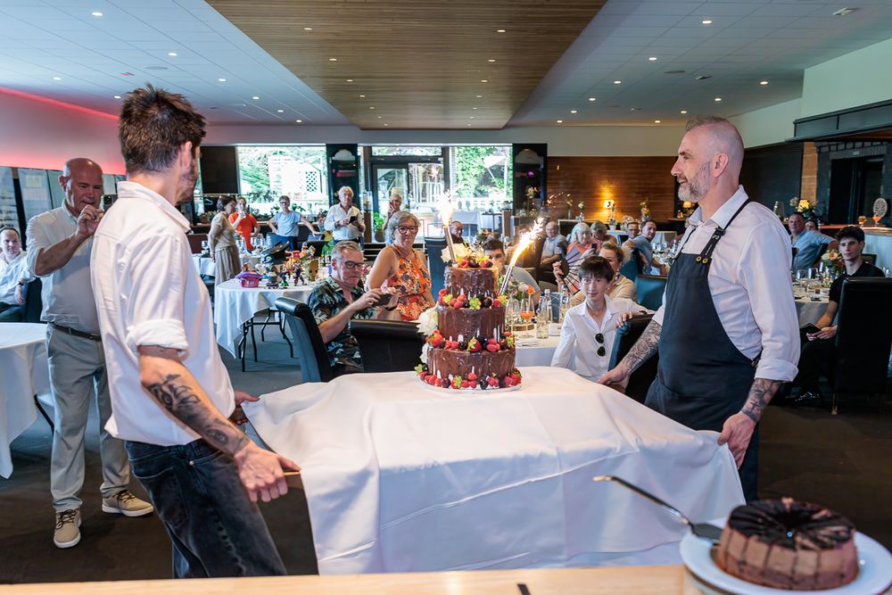 Two chefs present an elaborate celebration cake with sparklers at a festive indoor event filled with guests.