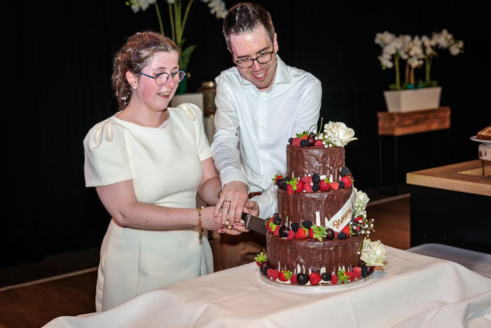 Bride and groom cutting a three-tier chocolate wedding cake decorated with fresh strawberries and flowers.