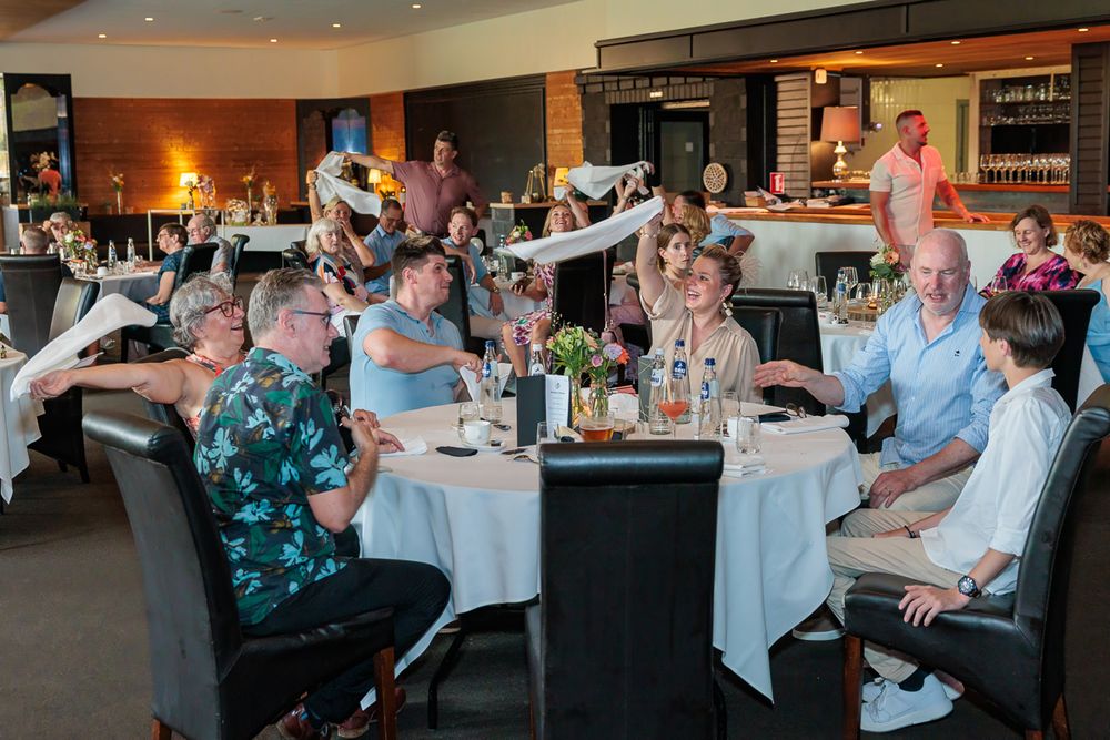 Group of people dining and celebrating at a restaurant, raising glasses and smiling around white round tables.