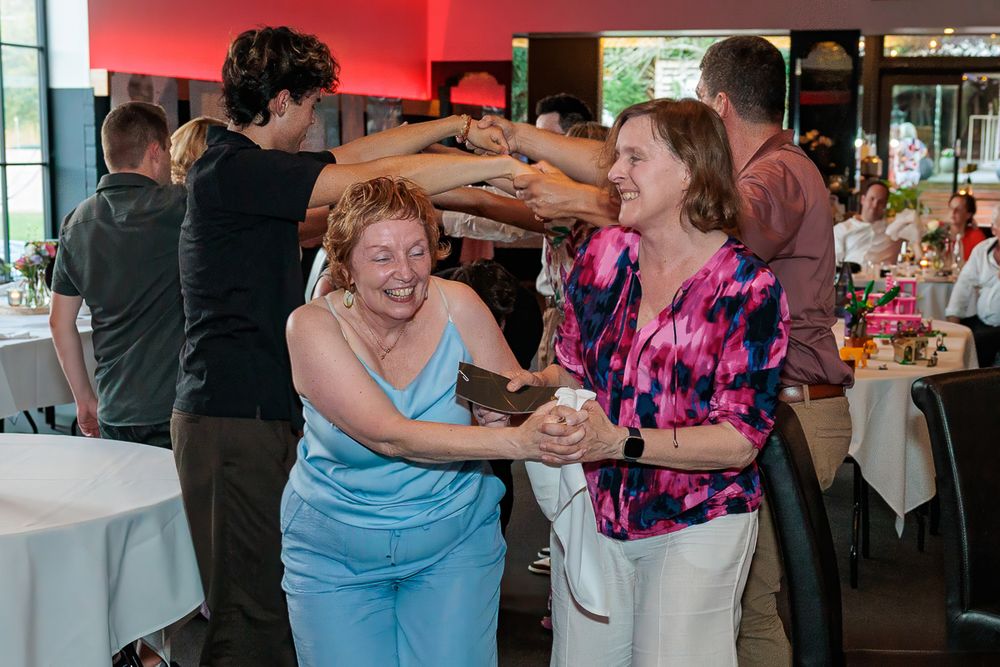 Two women laughing and dancing together at a festive indoor party event with guests dining in the background.