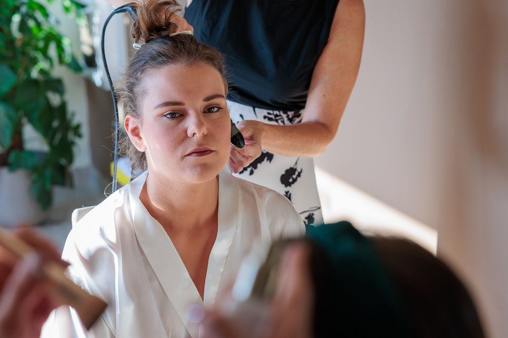 Bride in white robe getting hair and makeup done by stylists before her wedding ceremony.