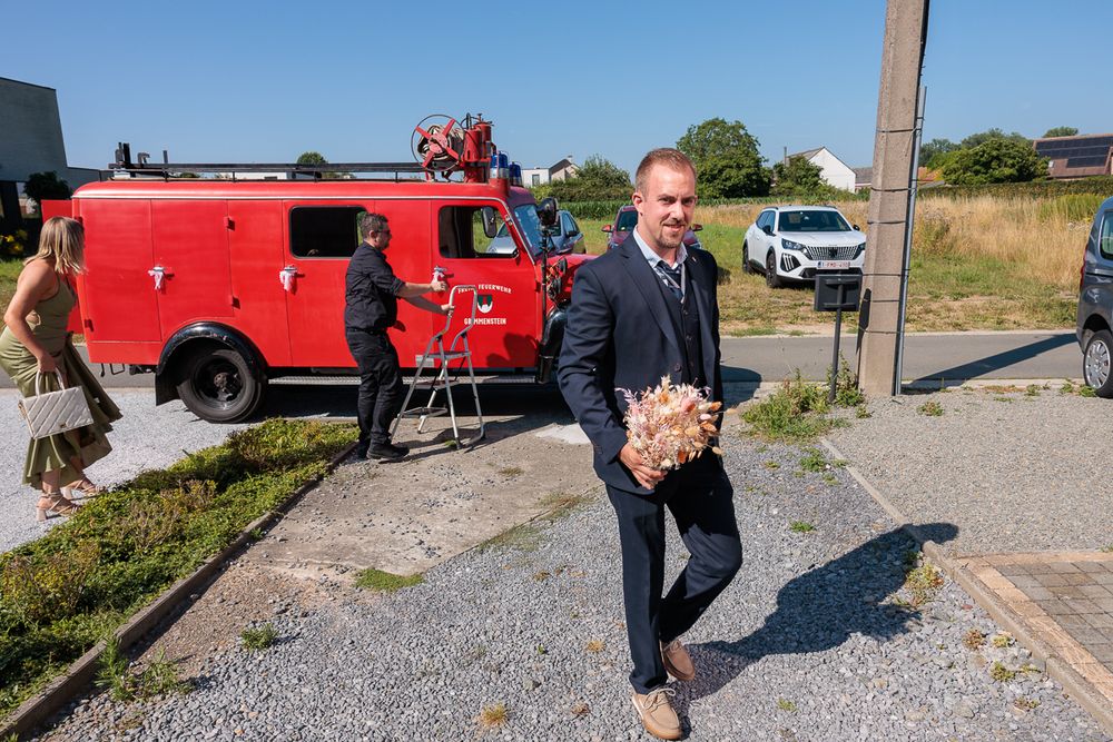 Man in blue suit carrying bouquet walks near vintage red fire truck on gravel lot on sunny day.