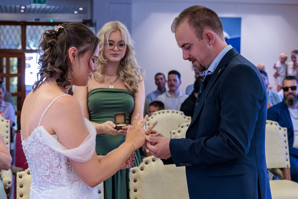 Bride placing wedding ring on groom's finger during ceremony, with bridesmaid in green dress watching in background.