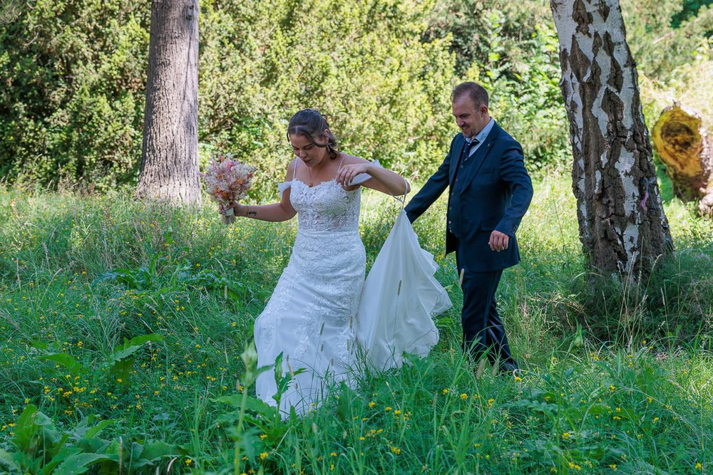A bride in a white gown holds a bouquet while a groom in a navy suit helps carry her train through a lush green meadow.
