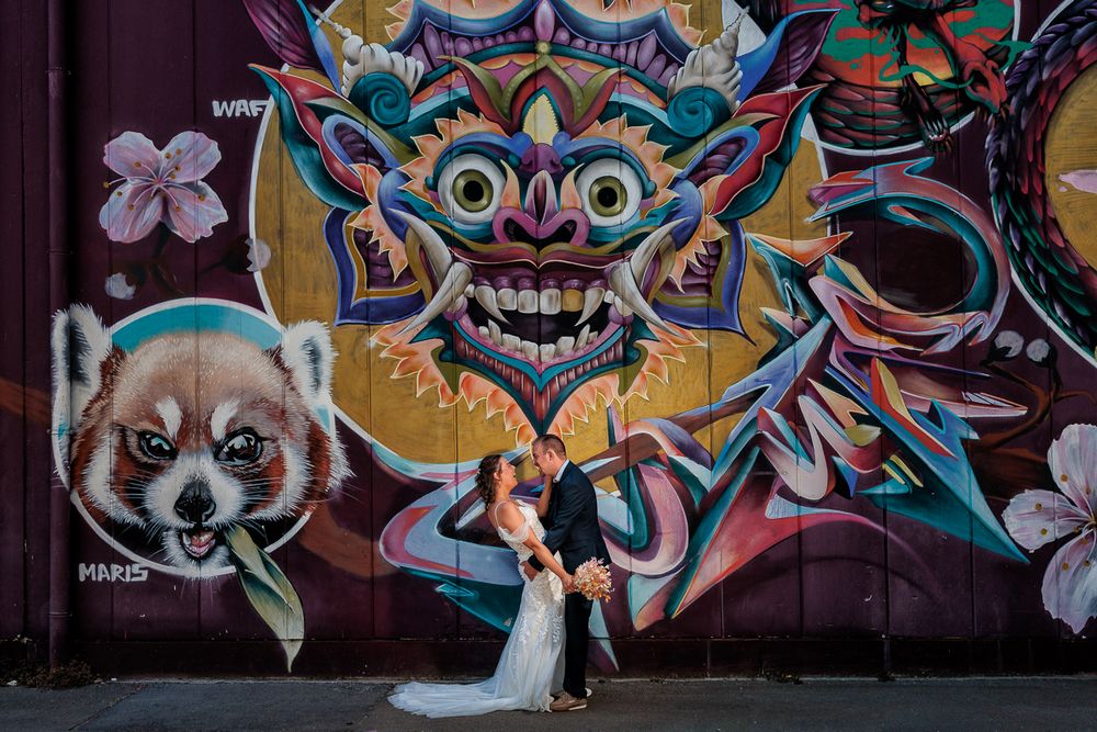 Bride and groom kissing in front of vibrant graffiti mural featuring a demon and red panda on a city wall.
