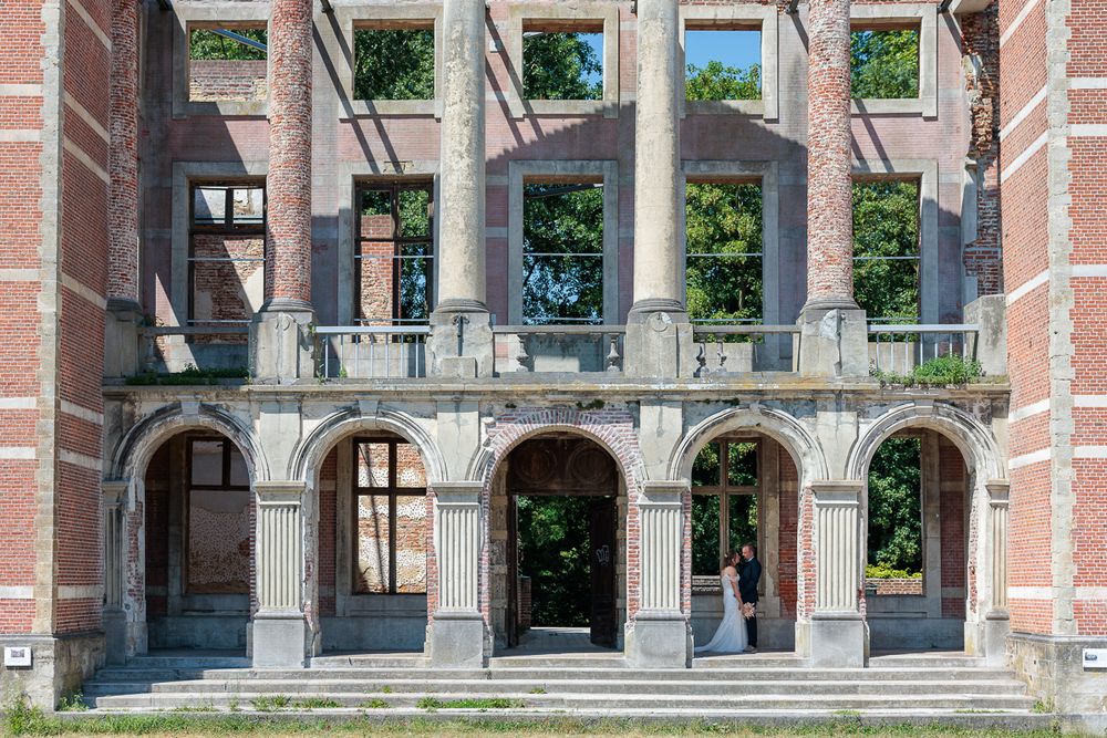 Abandoned red brick building with arched colonnades, balcony, and overgrown windows in a historic European architectural style.
