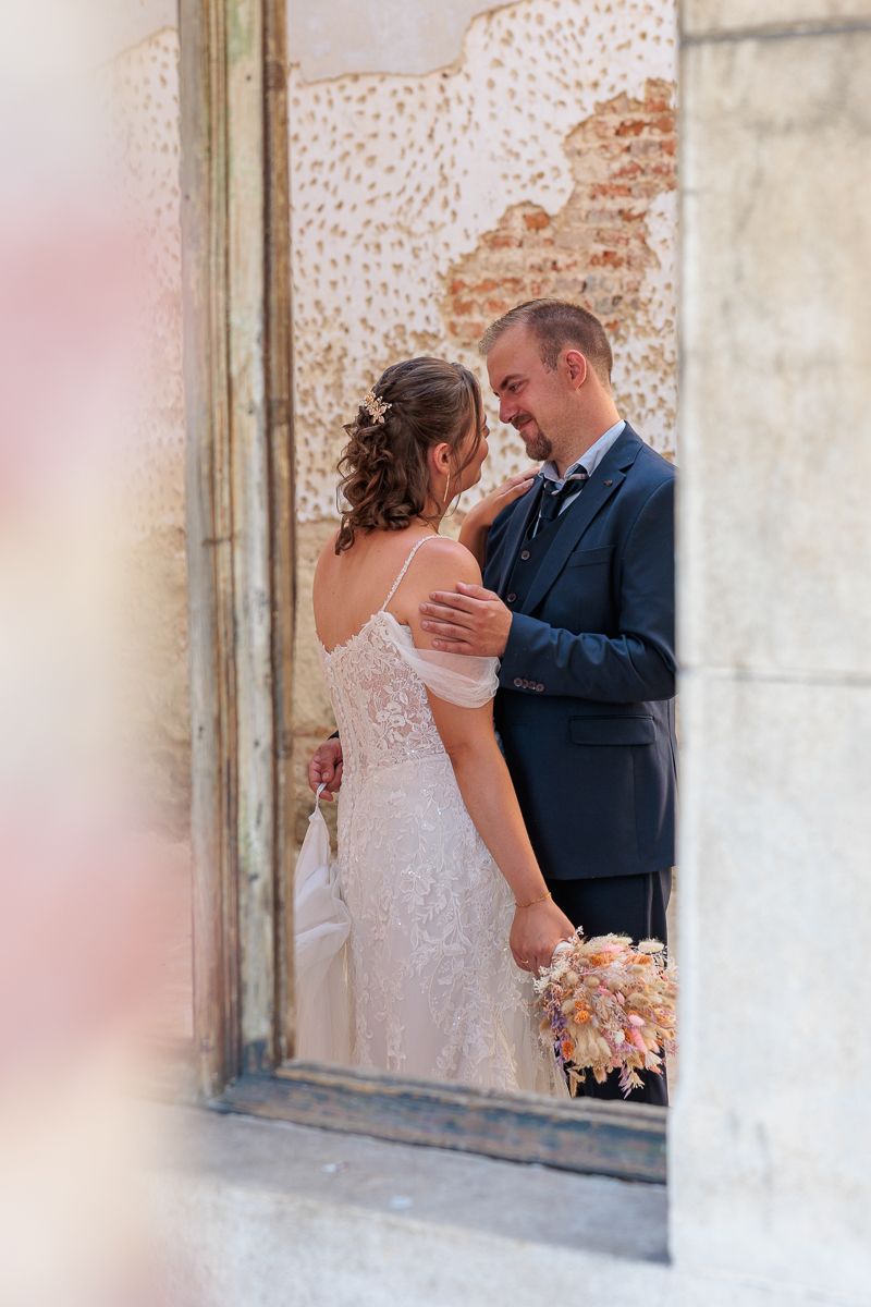 Bride and groom share an intimate moment in a rustic doorway, she in lace gown, he in navy suit holding bouquet.