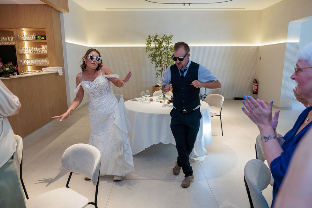Bride and groom dancing joyfully in sunglasses at their wedding reception while guests clap and celebrate.