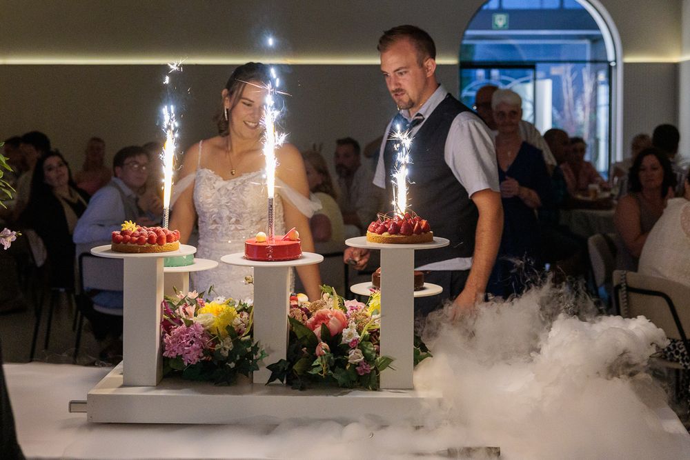 Bride and groom stand beside elegant wedding cakes with sparklers and dry ice fog at a romantic reception.