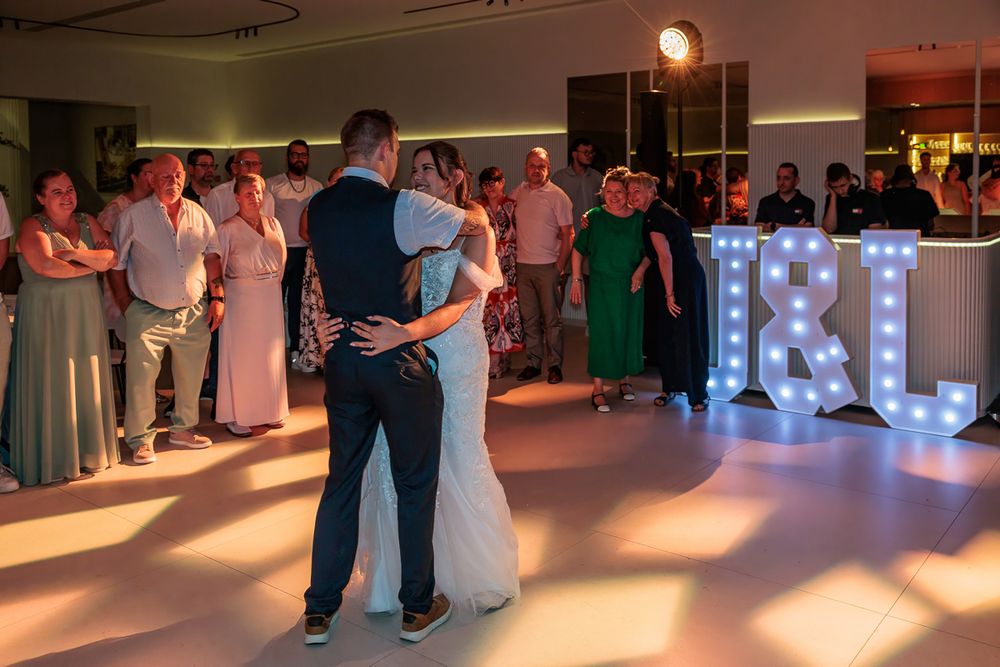 Bride and groom share first dance at wedding reception with illuminated I&L letter lights and guests watching.