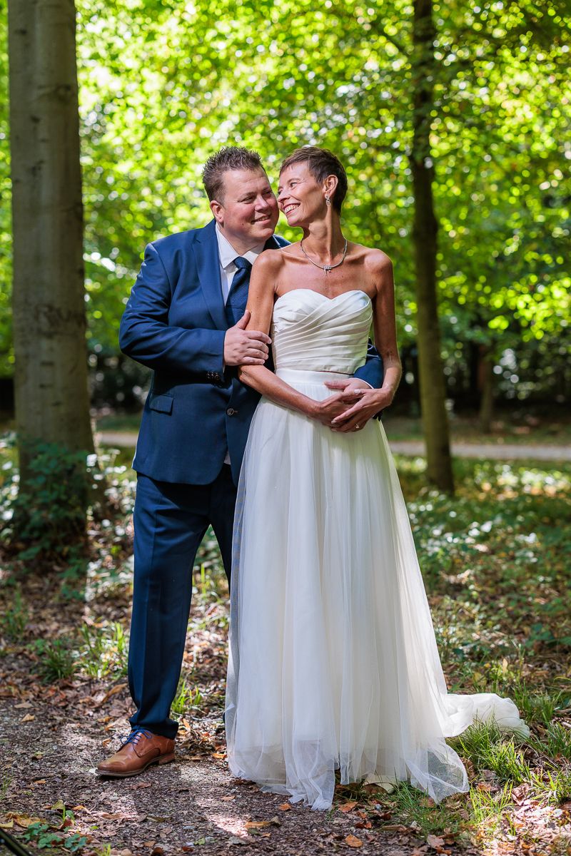 A bride in a white strapless gown and groom in a blue suit embrace romantically in a sunlit forest setting.