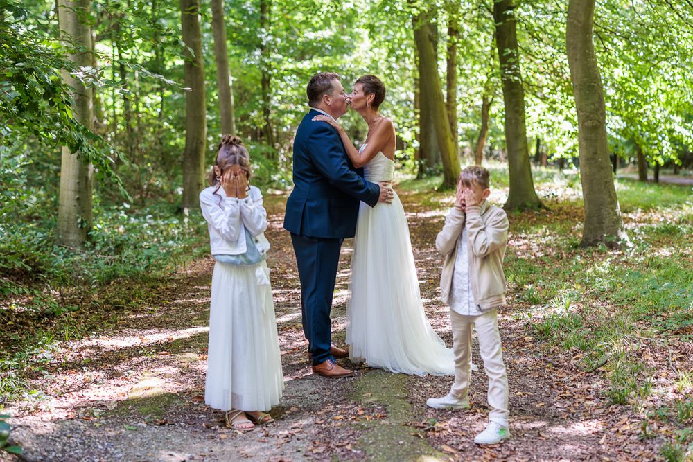 Bride and groom share a kiss in a forest while two children cover their eyes on their wedding day.