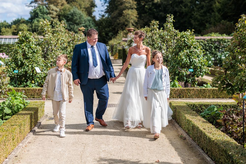 A bride and groom walk hand-in-hand with two children along a garden path on their sunny wedding day.