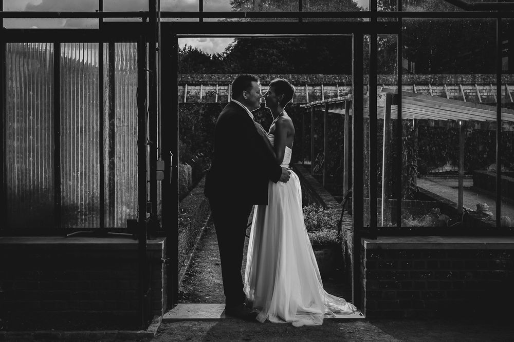 Black and white photo of a couple sharing a romantic kiss in a greenhouse doorway on their wedding day.