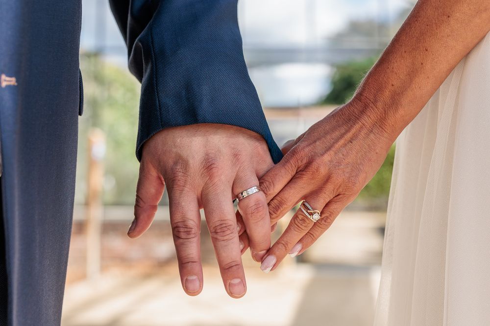 Newlyweds holding hands showing wedding rings, bride in white dress and groom in blue suit at outdoor wedding.