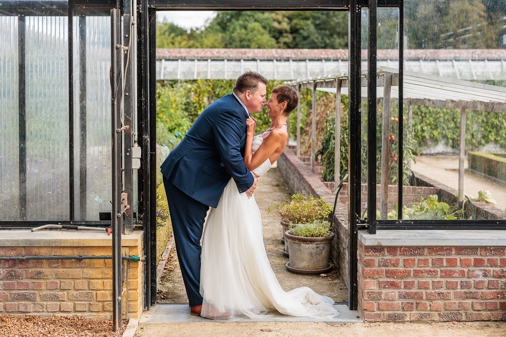 Bride and groom share a romantic kiss in a greenhouse doorway, bride in white gown, groom in navy suit.