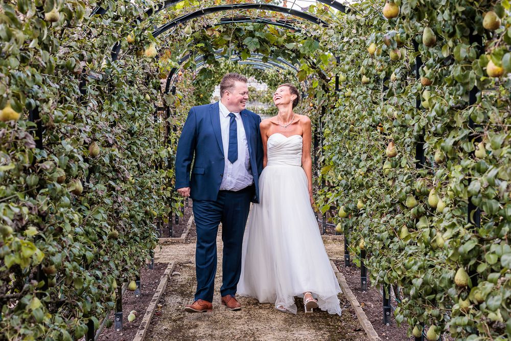 Bride and groom walking through a lush garden archway covered in greenery and fruit trees on their wedding day.