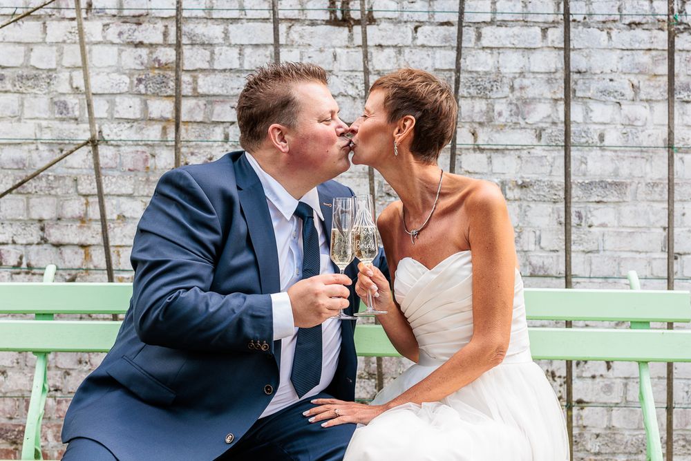 Newlywed couple kissing while holding champagne glasses, bride in white gown and groom in navy suit on green bench.