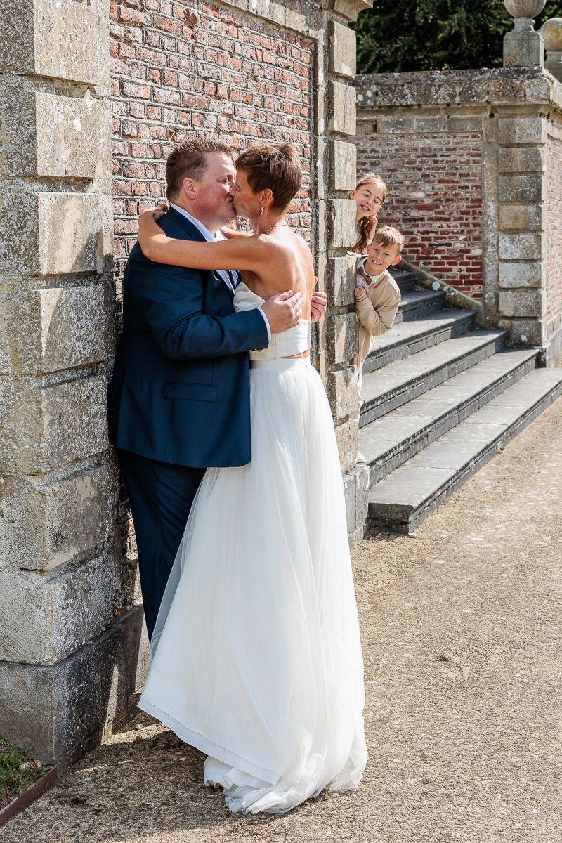 Bride and groom share a kiss against a stone wall while two children peek curiously from nearby steps.