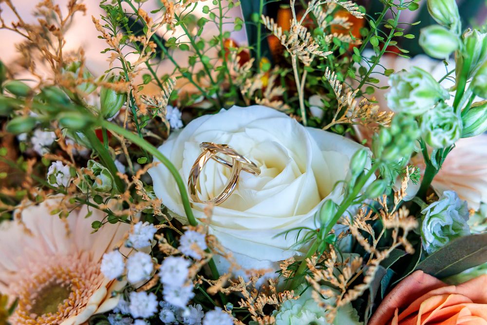 Two gold wedding rings resting on a white rose in a bridal bouquet with peach flowers and greenery.