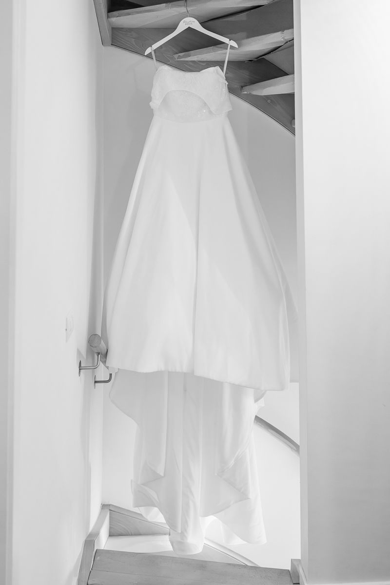 Black and white photo of an elegant white wedding dress hanging on a hanger in a stairwell doorway.