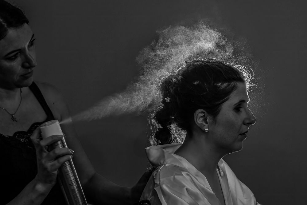 Black and white photo of a hairstylist spraying hairspray on a bride during wedding preparations.