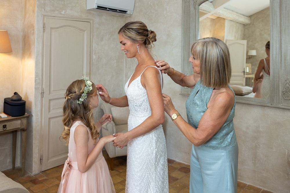 A bridal party helps prepare in a soft lit dressing room, adjusting details on a white lace wedding dress.