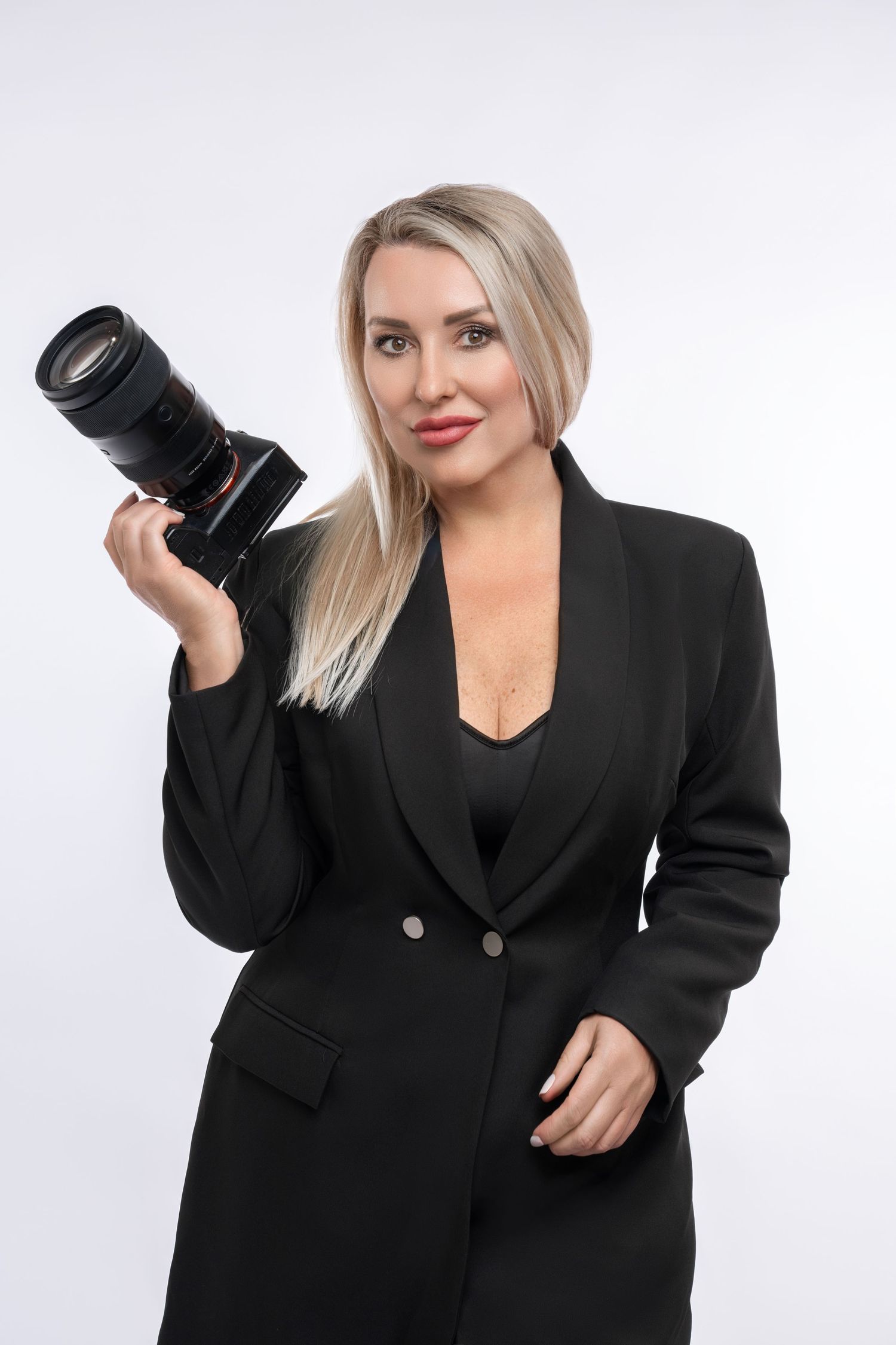 Blonde woman in black blazer holding a professional DSLR camera with telephoto lens on white background.