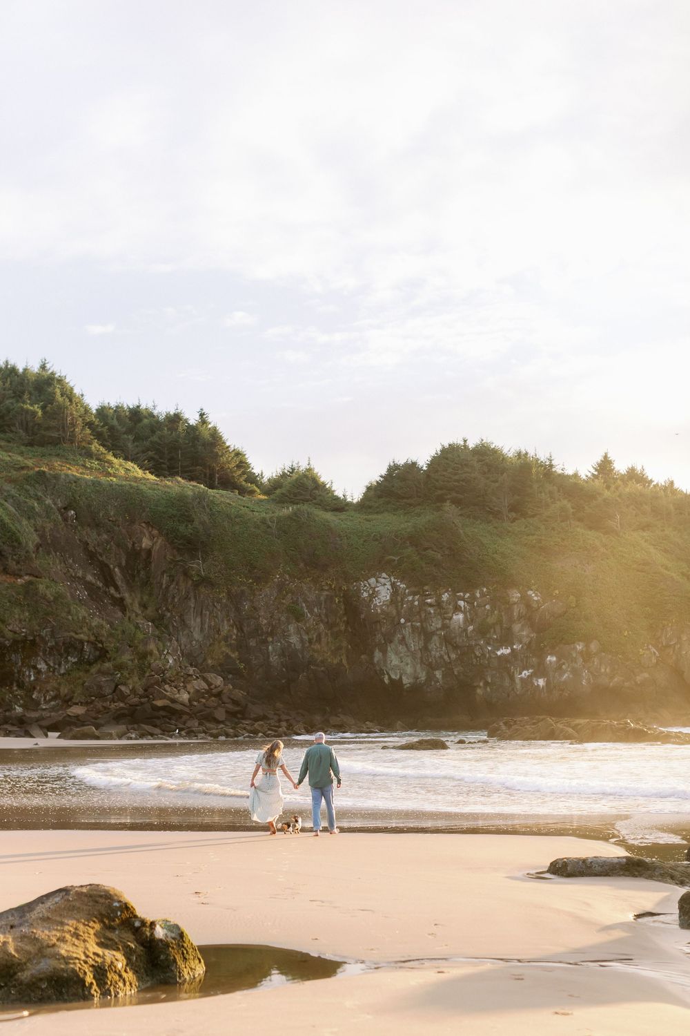 Couple holding hands walking on a sandy beach at golden hour with lush green cliffs and ocean waves in the background.