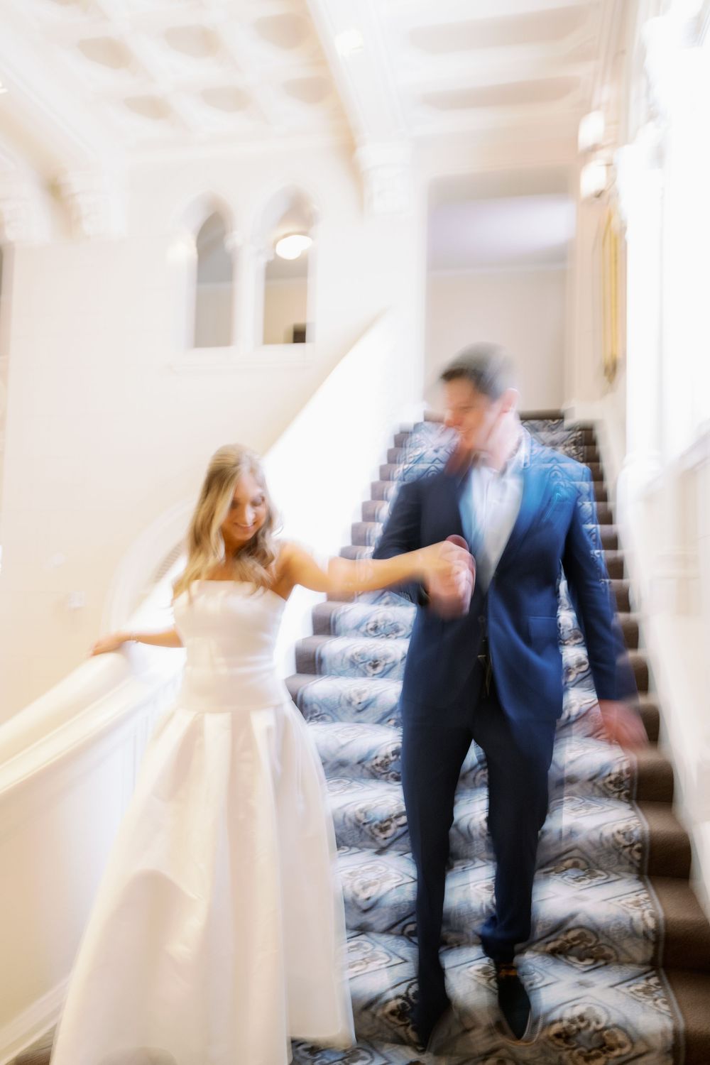 Two figures walking up a grand staircase with patterned carpet in a dreamy blurred motion-style wedding photo.