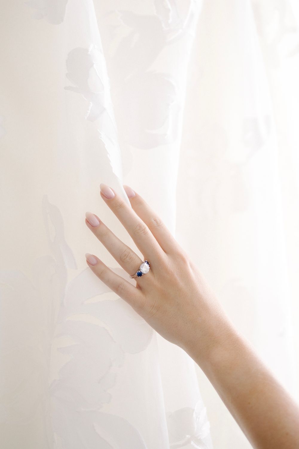 Elegant hand wearing a sapphire and pearl ring against a soft white floral curtain backdrop.
