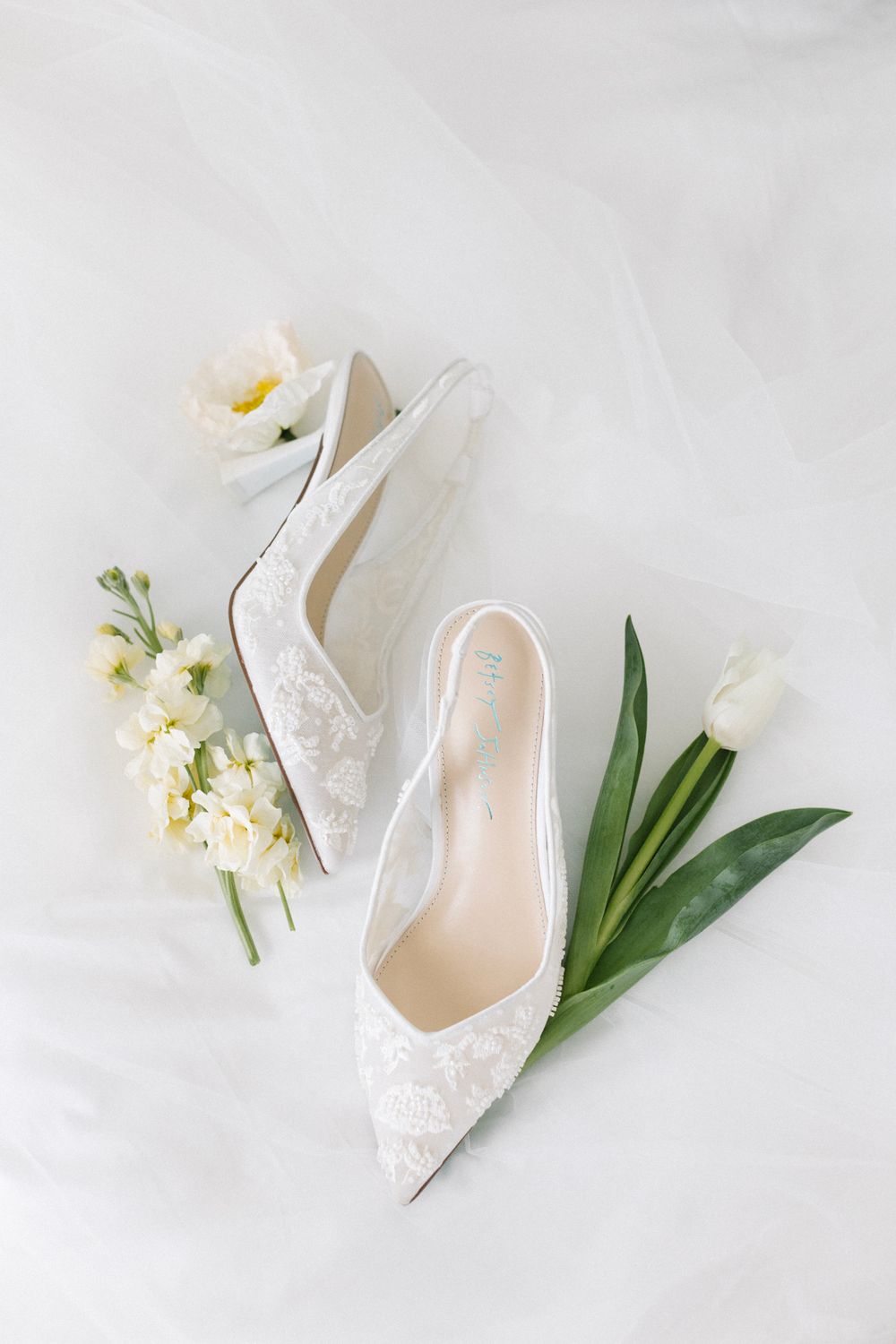 White lace bridal heels surrounded by fresh white tulips and flowers on a soft white fabric background.