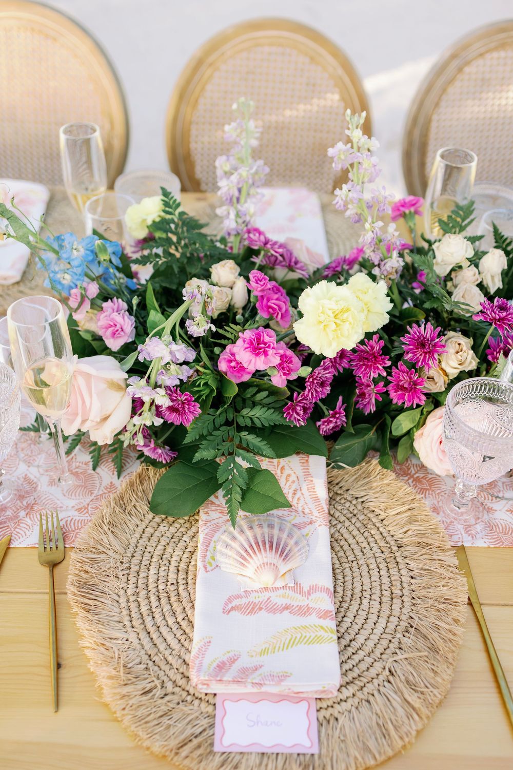 Elegant wedding table setting with vibrant floral centerpiece, woven charger plate, pink menu card, and gold cutlery.