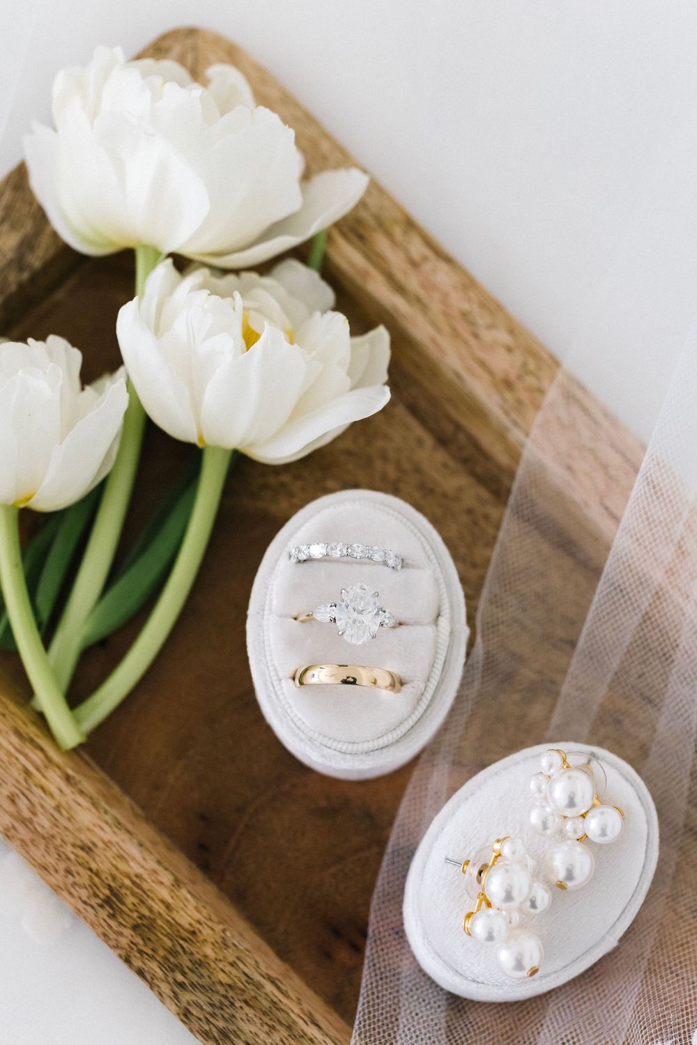 Elegant wedding rings displayed in a white velvet box beside white tulips on a wooden tray with pearl accessories.