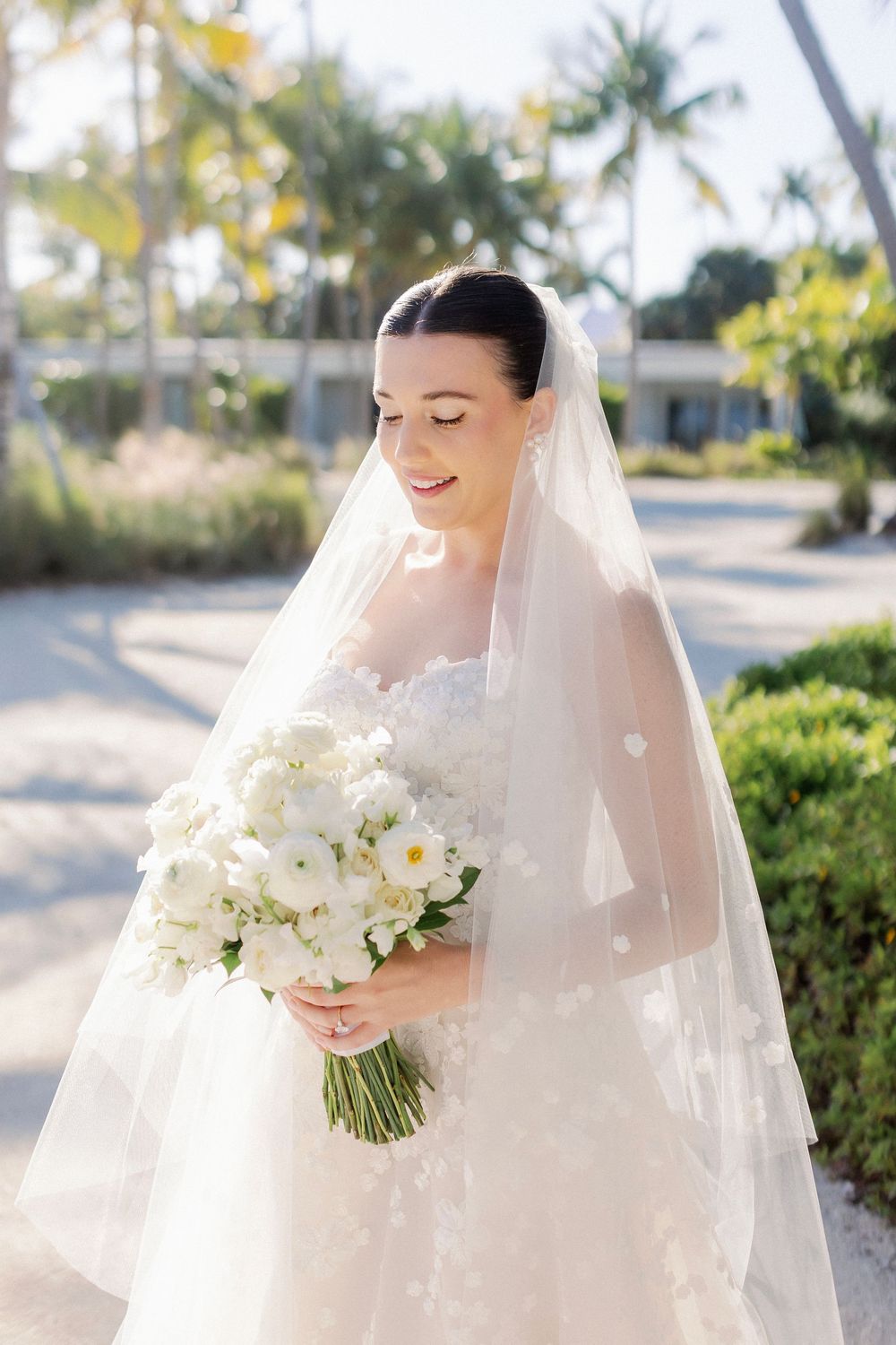 Smiling bride in white lace gown and veil holds white floral bouquet outdoors with palm trees.