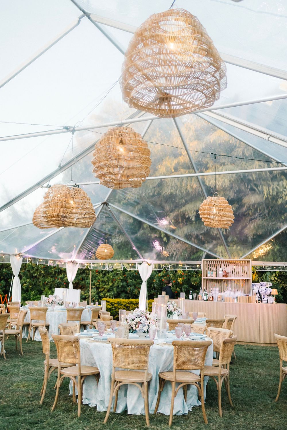 Elegant outdoor wedding reception under clear tent with wicker pendant lights, round tables, and wooden chairs on green lawn.