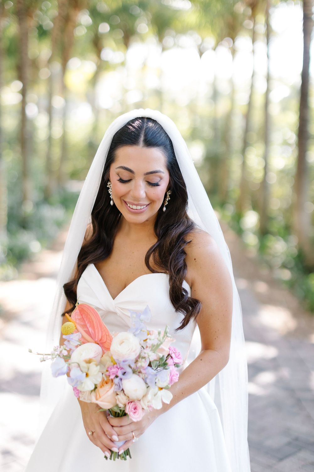 Smiling bride in strapless white gown and veil holds colorful bouquet of pastel flowers outdoors.
